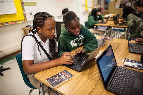 两个高中女生在教室里共用一台笔记本电脑。 其中一个身穿白色 Polo 衫的人正聚焦在屏幕上。 她的朋友穿着绿色运动衫，对着屏幕微笑。